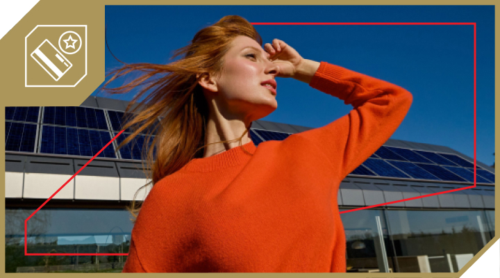 Na sliki je dekle z dolgimi lasmi, oblečena v oranžen pulover, ki stoji na prostem pred stavbo s sončnimi celicami na strehi. Pogled ima usmerjen proti soncu, v ozadju pa je jasno modro nebo.