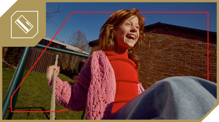 Na sliki je nasmejana deklica z dolgimi lasmi, oblečena v oranžen pulover, ki se ziblje na gugalnici na dvorišču. V ozadju sta opečnata hiša in modro nebo.