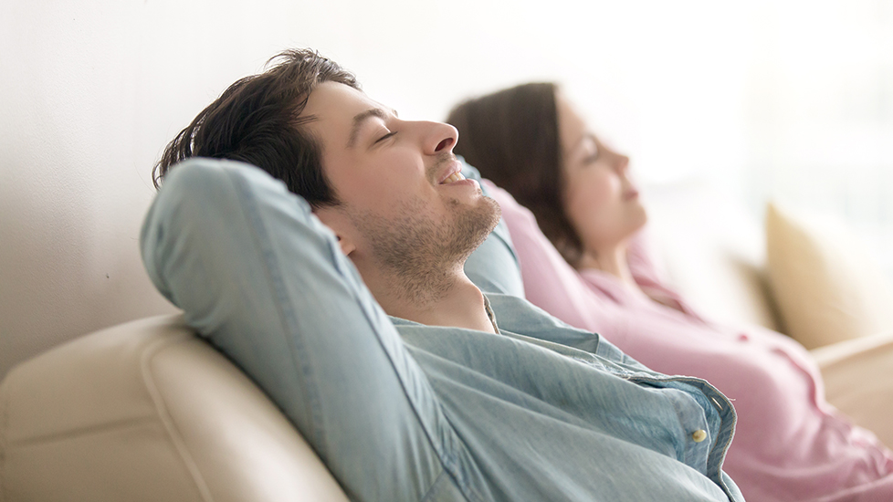 Na sliki sta moški in ženska, ki sproščeno sedita na kavču, z naslonjenimi glavami in zaprtimi očmi, vidno uživata v udobju in toplini doma.
