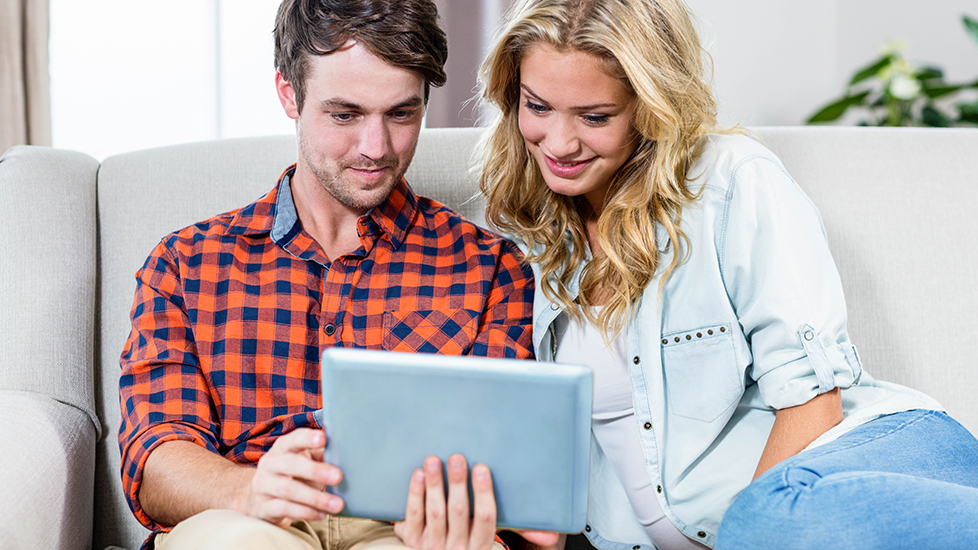 Na sliki sedita moški in ženska na kavču ter skupaj gledata v tablico, ob tem pa se nasmihata.