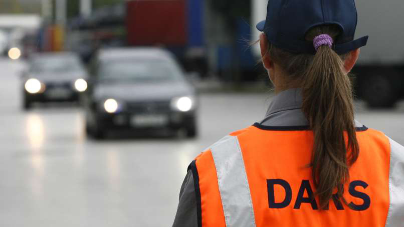 Na sliki je oseba v odsevnem oranžnem jopiču z napisom 'DARS' na hrbtu. Oseba stoji ob cesti, kjer se približujejo avtomobili.