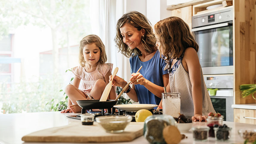 Na sliki je mati z dvema hčerkama v sodobni kuhinji. Skupaj pripravljajo obrok na štedilniku, obkrožene z živili in pripomočki za kuhanje, medtem ko se smehljajo in uživajo v skupnem času.