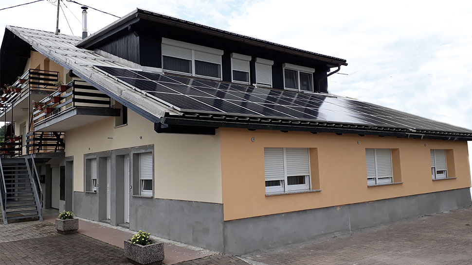 Na sliki je večja dvonadstropna hiša z rumeno-bež fasado in sivo osnovo. Na strehi je nameščena velika sončna elektrarna, ki pokriva skoraj celotno strešno površino. Na levi strani hiše je zunanje kovinsko stopnišče, ki vodi do nadstropja z balkonom. Balkoni so okrašeni z rdečimi cvetličnimi koriti. Spodaj so bela okna z roletami, pred hišo pa betonsko dvorišče z nekaj cvetličnimi koriti s cvetjem. Nebo v ozadju je oblačno.