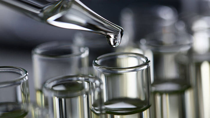 Na sliki je prikazano kapljanje tekočine iz pipete v steklene epruvete v laboratoriju.