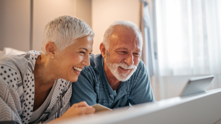 Na sliki sta nasmejana starejši moški in ženska, ki skupaj gledata v tablico. Sedita v prijetnem, svetlem prostoru in delujeta zadovoljno ter sproščeno.