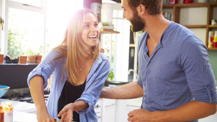 Na sliki sta mlajši moški in ženska v svetli kuhinji. Med kuhanjem se smejita in pogovarjata, kar ustvarja občutek sproščenosti in topline domačega vzdušja.