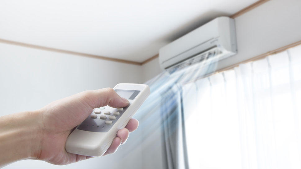 Na sliki je notranja enota klimatske naprave, nameščena na steni ob oknu z belimi zavesami. Iz naprave piha hladen zrak, ki je ponazorjen z modrimi žarki. V ospredju je vidna roka, ki drži bel daljinski upravljalnik in ga usmerja proti napravi.