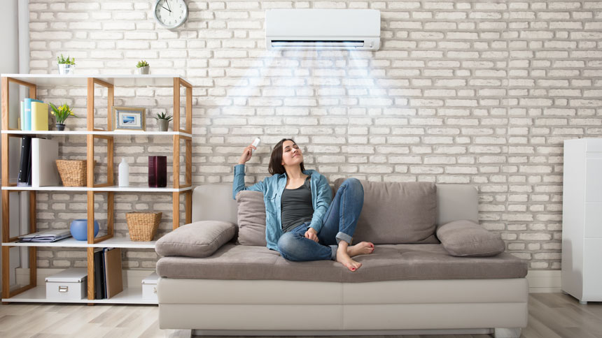 Na sliki je ženska, ki sproščeno sedi na sivi sedežni garnituri v dnevni sobi. V roki drži daljinski upravljalnik in uživa v svežem zraku, ki ga piha klimatska naprava, nameščena na steni nad njo. Na steni je vidna bela opečna dekoracija, poleg sedežne garniture stoji bela omara, na levi strani prostora pa knjižna polica z različnimi predmeti in rastlinami. Nad klimatsko napravo visi okrogla stenska ura.