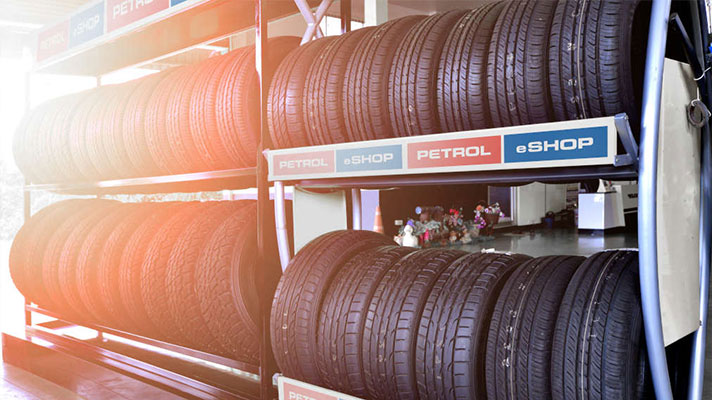 Na sliki so avtomobilske gume zložene na kovinskih policah v trgovini. Nad gumami je napis Petrol eSHOP.