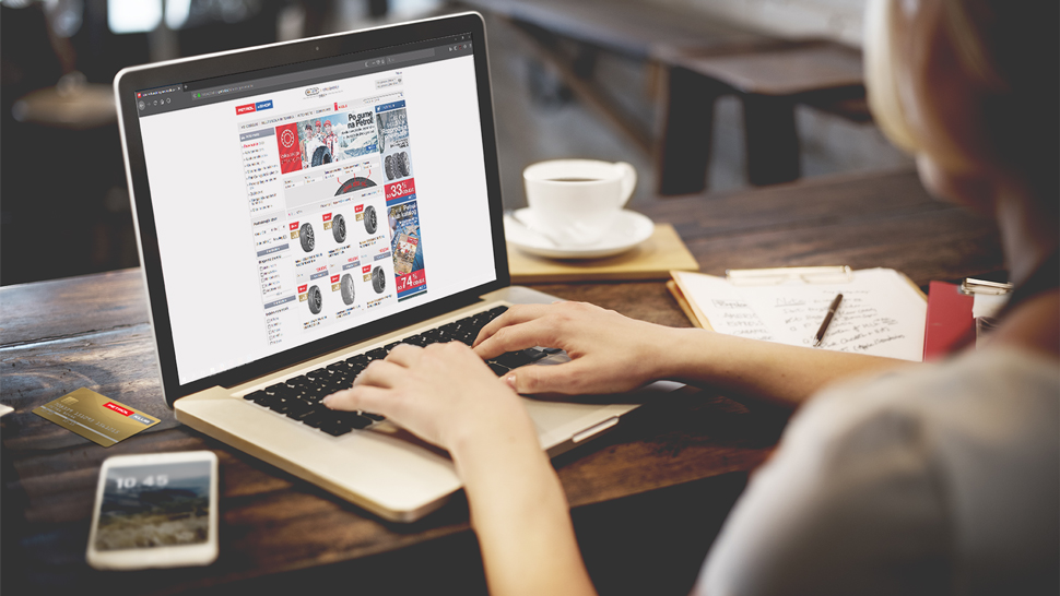 Na sliki oseba uporablja prenosni računalnik in brska po spletni trgovini z avtomobilskimi gumami. Na mizi so poleg računalnika še mobilni telefon, beležka, kava in kreditna kartica.