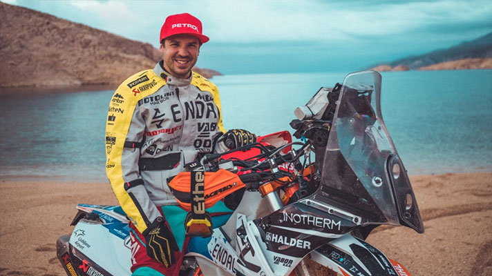 Na sliki je motorist v tekmovalni opremi, ki sedi na motorju ob jezeru. Nosi rdečo kapo Petrol in rumeno jakno s sponzorskimi napisi.