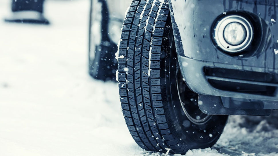 Na sliki je sprednji del avtomobila z žarometom, v ospredju pa pnevmatika z zimskim profilom. Avto stoji na zasneženi podlagi na kateri so vidne snežinke.