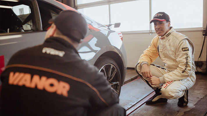 Na sliki sta mehanik in dirkač ob avtomobilu v delavnici. Dirkač v kombinezonu in s kapo se pogovarja z mehanikom, ki nosi jakno z napisom Vianor.