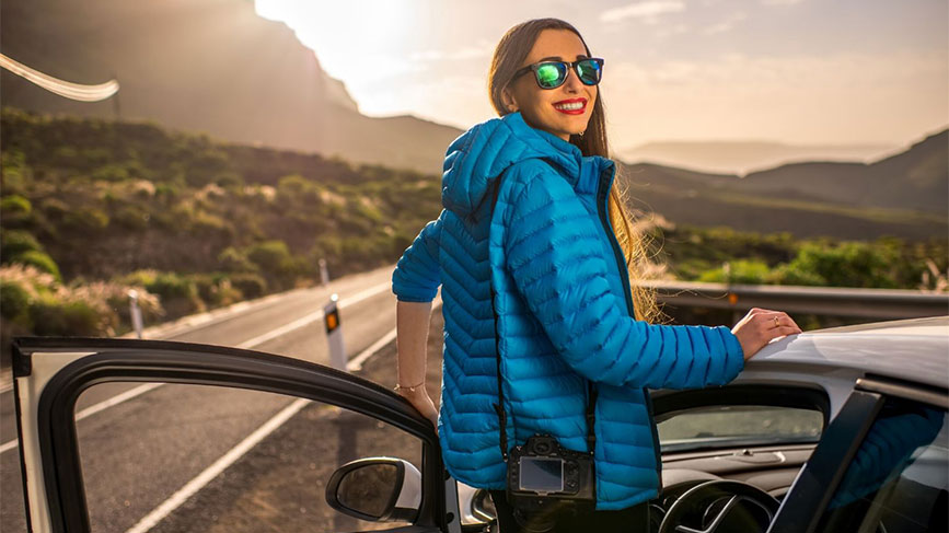 Na fotografiji je dekle v modri puhasti jakni in sončnih očalih, ki stoji ob odprtih vratih avtomobila. Nasmejana se ozira proti soncu, v ozadju pa se razprostira cesta in slikovita pokrajina, obsijana s toplimi žarki.