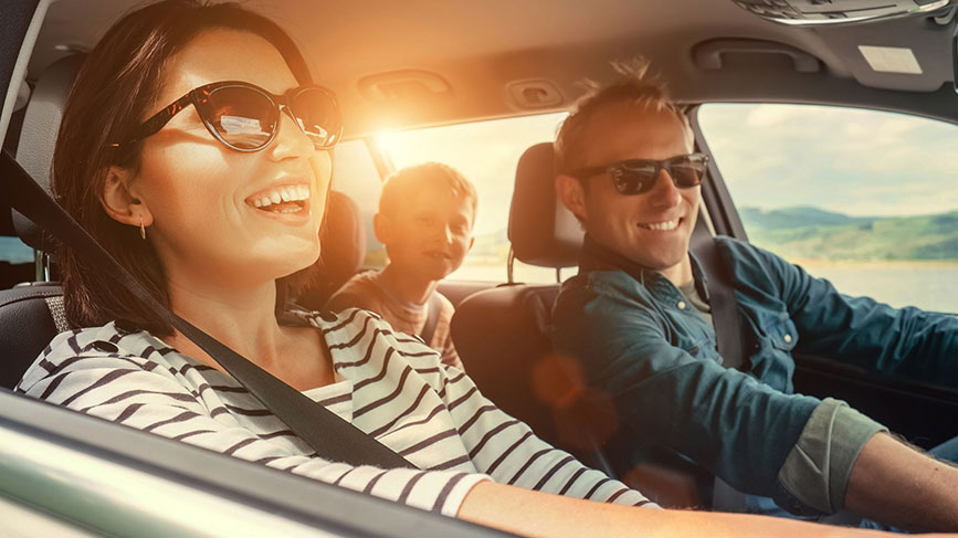 Na sliki je družina v avtomobilu – moški vozi, ženska sedi na sovoznikovem sedežu, zadaj pa je otrok. Vsi so nasmejani, sproščeni in nosijo sončna očala. Sončni žarki, ki padajo skozi okno, dajejo prizoru občutek poletja in brezskrbnega potovanja.