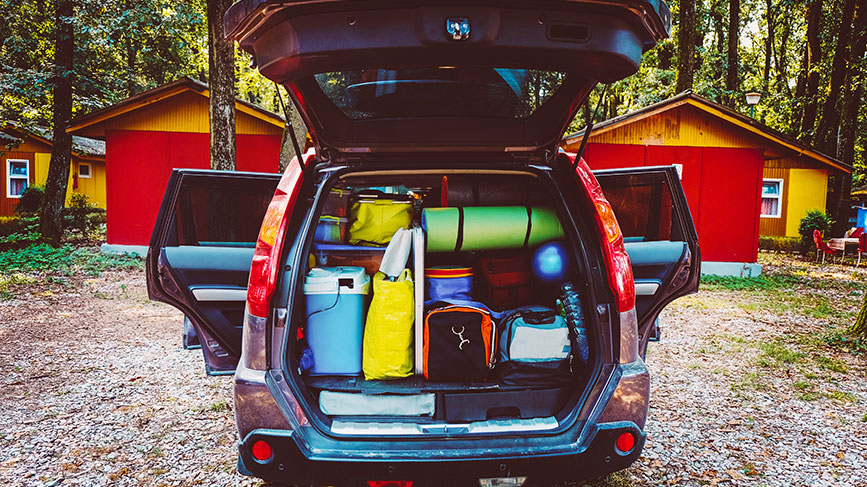 Na sliki je odprt prtljažnik avtomobila, natančno napolnjen s potovalno opremo – torbami, hladilno skrinjo, spalnimi vrečami in podlogami za kampiranje. Vozilo stoji na dvorišču med rdečimi in rumenimi lesenimi hiškami v gozdu.