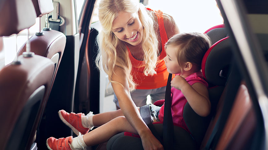 Na fotografiji je ženska, ki pomaga otroku, da se pripne v otroški avtosedež na zadnjem sedežu avtomobila. Ženska se smehlja, otrok pa mirno sedi v sedežu, medtem ko mu zapenja varnostni pas. Avtosedež je črn z rdečimi obrobami.