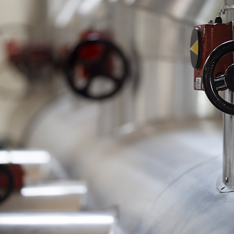 slika prikazuje detajl industrijske instalacije: najbližje kameri je kovinski ventil s črnim krožnim ročajem in rdečo osrednjo zardevelo ročko; v ozadju so zamegljene kovinske cevi in drugi ventili.