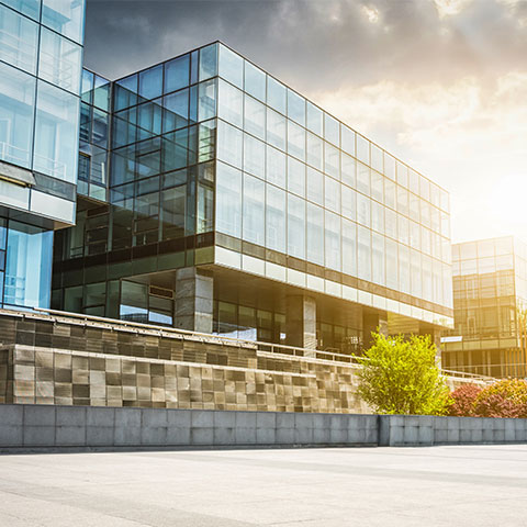 Slika prikazuje sodobno poslovno stavbo s stekleno fasado, kamnitim zidom in sončno svetlobo v ozadju.