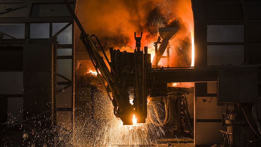 Na sliki je prikazan industrijski proces taljenja kovine v peči, iz katere izhajajo iskre in žareča staljena kovina.