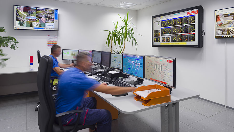 Na sliki je prikazan nadzorni center z dvema zaposlenima, ki spremljata več računalniških zaslonov in prikazovalnikov v nadzorni sobi