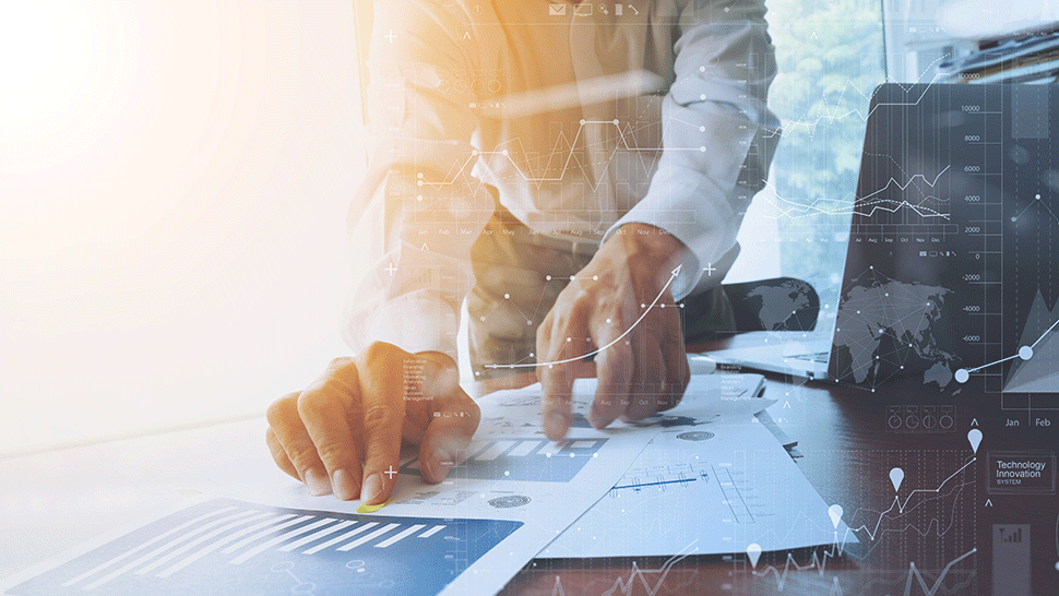 Slika prikazuje poslovneža, ki pregleduje dokumente z grafikoni in analizami na mizi.