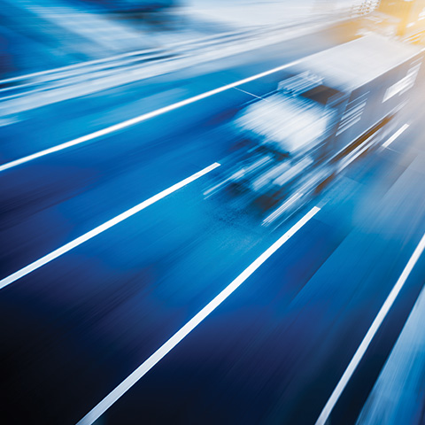Dinamična fotografija iz zraka avtomobila ali tovornjaka, ki se giblje po cestni površini, prikazana v zamrznjenem posnetku z zamegljenimi horizontalnimi svetlobnimi sledmi – poudarek je na hitrosti in mobilnosti.
