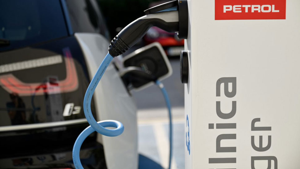 Na sliki je prikazana polnilna postaja Petrol za električna vozila, na kateri je priključen kabel modre barve, v ozadju pa je parkiran električni avtomobil.