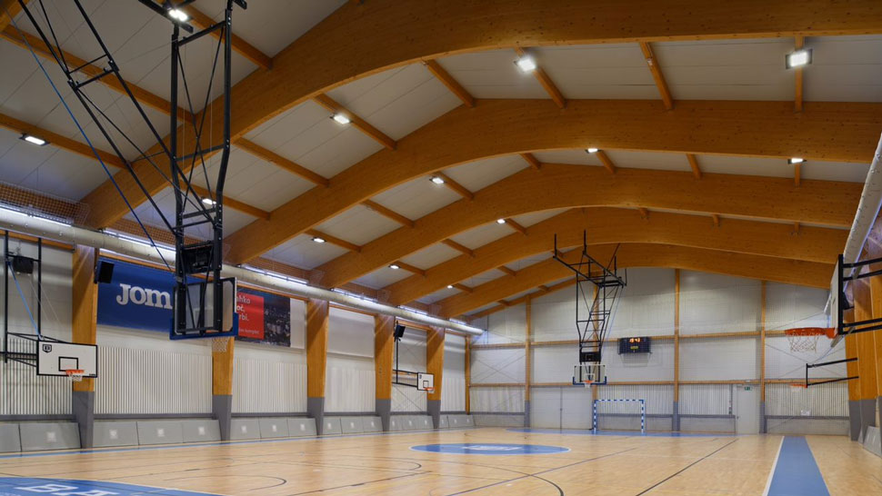 Na sliki je prikazana notranjost sodobne športne dvorane z lesenimi loki, košarkarskimi koši in označenim igriščem.