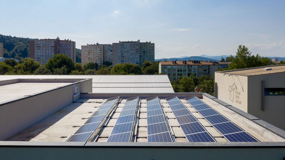 Na sliki je prikazana streha stavbe s sončnimi celicami, ob kateri je stavba z napisom 'Šolski center Ljubljana'; v ozadju so drevesa, večstanovanjski bloki in hribovita pokrajina.