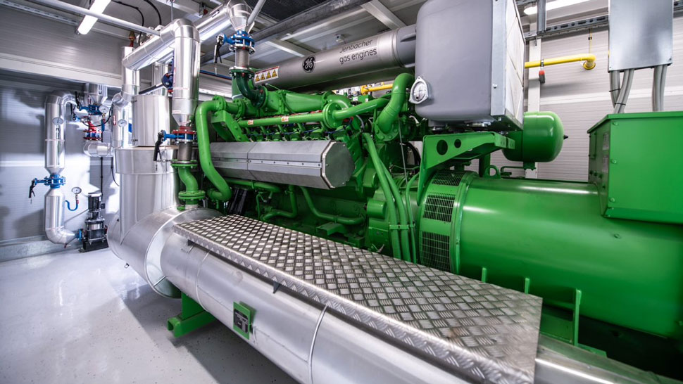 Na sliki je prikazan velik industrijski plinski motor zelene barve z označbo 'gas engines'; obdan je s številnimi cevmi, ventili in kovinsko delovno platformo s protizdrsno površino, vse skupaj nameščeno v svetli strojnici.