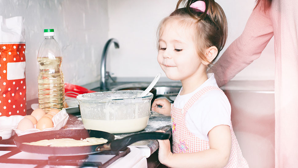 Na sliki je deklica v kuhinji, ki stoji ob štedilniku, kjer se v ponvi peče palačinka. Na pultu so skleda s testom, jajca, steklenica olja in moka. Deklica ima nasmešek na obrazu, ob njej pa je prisotna tudi odrasla oseba.