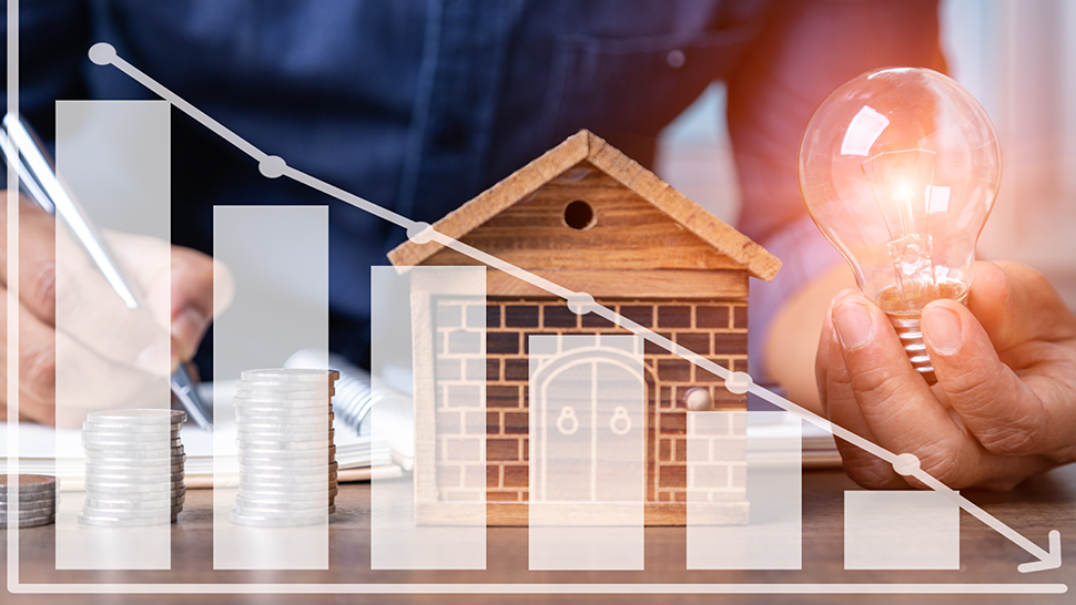 Na sliki je prikazana simbolična upodobitev varčevanja z energijo: oseba drži prižgano žarnico poleg makete hiše, na mizi so naloženi kovanci, preko slike pa je narisan graf z padajočim trendom stroškov.