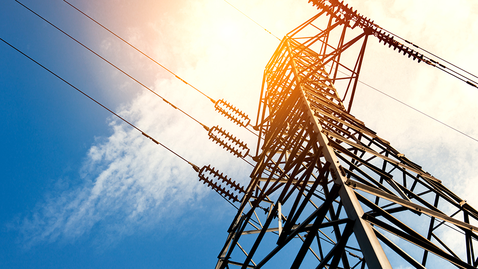 Na sliki je jekleni daljnovod z električnimi vodi, ki se dviguje proti nebu. Pogled je usmerjen navzgor, sončni žarki pa osvetljujejo konstrukcijo in dajejo prizoru toplo svetlobo. V ozadju je jasno nebo z nekaj oblački.