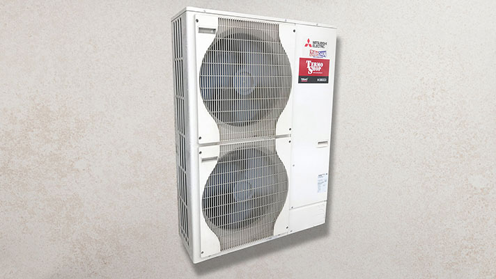 Na sliki je zunanja enota toplotne črpalke Mitsubishi Electric. Enota je bele barve in ima dva velika ventilatorja, nameščena en nad drugim, ki sta zaščitena z rešetko. Na zgornjem desnem delu naprave je logotip Mitsubishi Electric in nalepka z oznako »Zubadan« ter dodatnimi podatki. Enota je postavljena ob svetlo sivo steno.