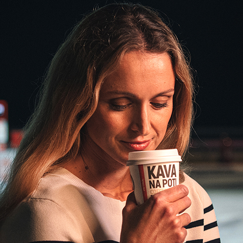 Dekle v črtasti majici drži skodelico kave z napisom 'Kava na poti' in uživa v toplem napitku.