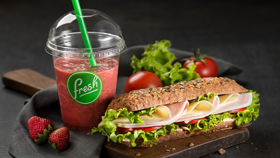 Petrol Fresh ponudba s sendvičem polnim šunke, sira in solate ter osvežilnim smoothiejem z jagodami v prozorni embalaži z zeleno slamico.
