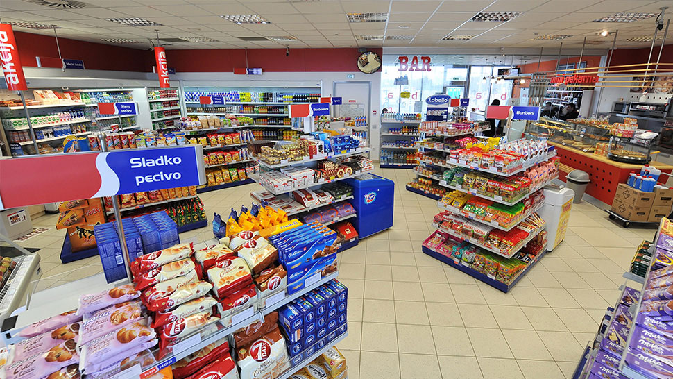 Notranjost trgovine na bencinskem servisu Petrol z raznoliko ponudbo izdelkov, med drugim sladkim pecivom, prigrizki, pijačami in osnovnimi živili, razporejenimi po policah in hladilnikih.