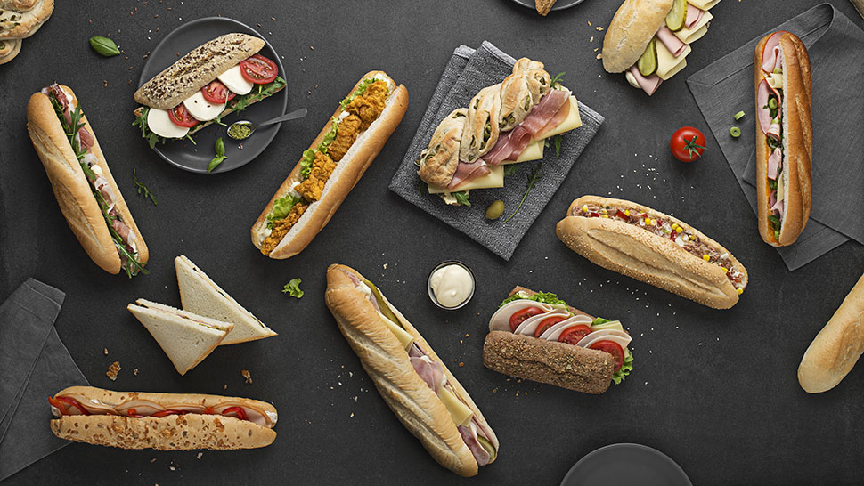 Raznolika ponudba sendvičev, baget in toastov, estetsko razporejenih na temnem ozadju s pogrinjki in dodatki.