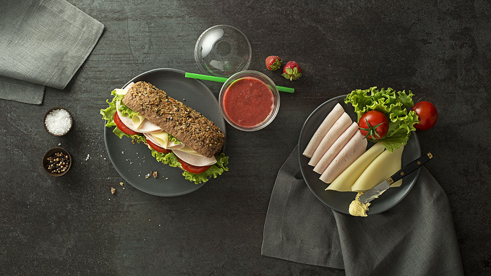 Polnozrnat sendvič s šunko, sirom, solato in paradižnikom na krožniku, ob njem kozarec jagodnega napitka ter krožnik z rezinami šunke, sira, solato in paradižnikom, vse postavljeno na temnem ozadju.