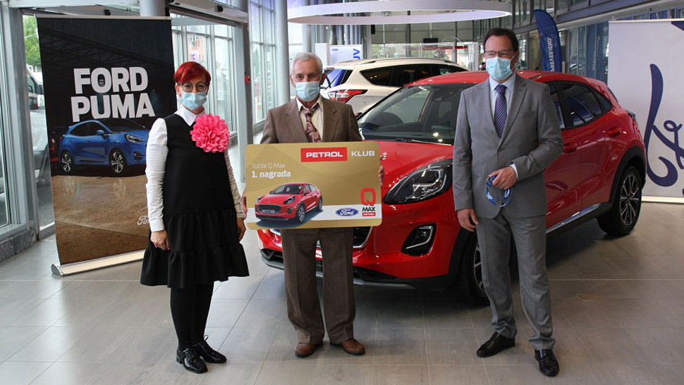 Trije ljudje z zaščitnimi maskami stojijo v salonu ob rdečem avtomobilu Ford Puma. Srednji moški drži veliko simbolično Petrol klub kartico z napisom '1. nagrada'. Ob strani je razstavni pano z napisom Ford Puma.