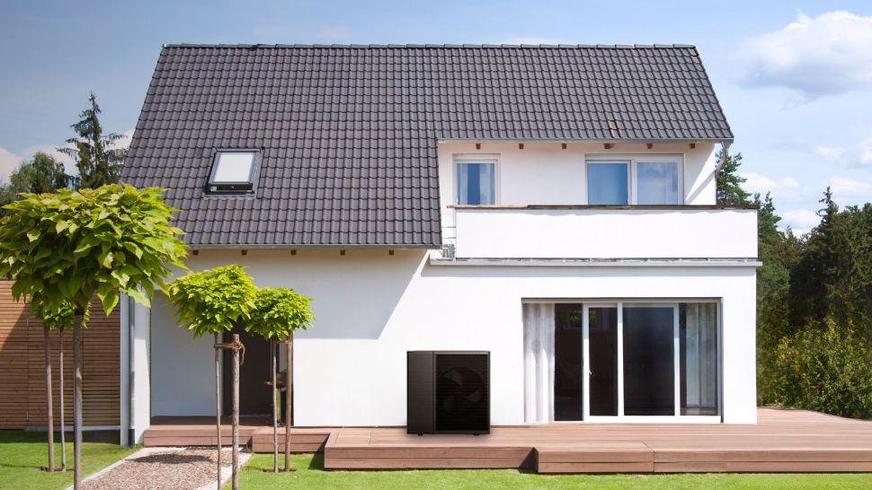 Na sliki je moderna dvonadstropna hiša z velikimi steklenimi vrati in balkonom. Streha je temno sive barve s strešnim oknom. Pred hišo je lesena terasa, ob njej pa urejena trata in mlada drevesa z bujnimi krošnjami. Ob steni hiše na terasi stoji toplotna črpalka pravokotne oblike v črni barvi. V ozadju se vidi zelenje in modro nebo z nekaj oblaki.