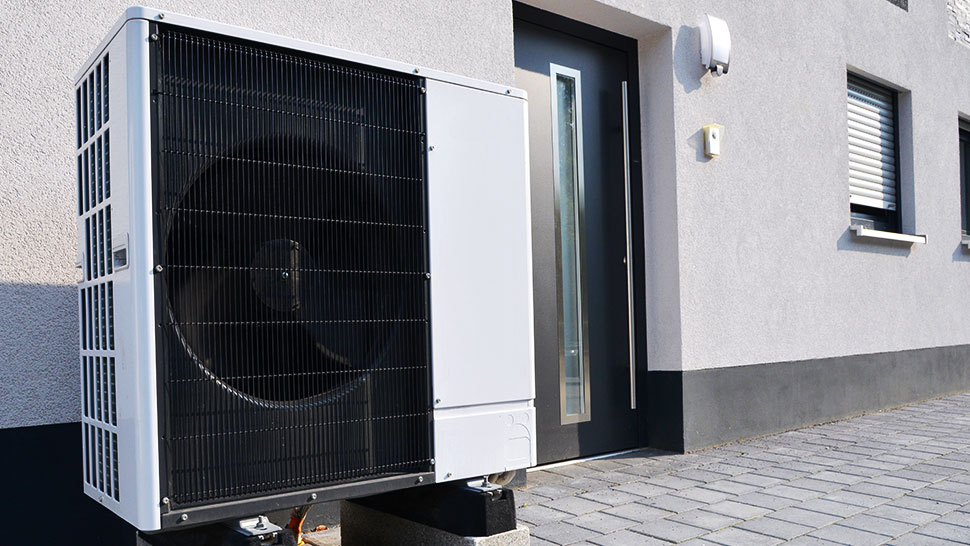 Na sliki je zunanja enota toplotne črpalke, ki je nameščena ob zunanji steni stanovanjske hiše. Enota je bela, s črno rešetko na sprednji strani, skozi katero je viden velik ventilator. Postavljena je na betonski podstavek tik ob vhodnih vratih hiše. Hiša ima sivo fasado, okna z belimi roletami in temno vhodno vrata z dolgim ozkim steklenim delom. Pred enoto je tlakovan prostor iz betonskih plošč.