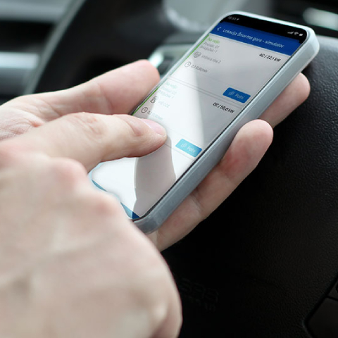 Na sliki je roka osebe, ki v avtomobilu uporablja pametni telefon z odprto aplikacijo OneCharge. Na zaslonu so prikazane informacije o času, količini energije in stanju polnjenja. V ozadju je volan vozila.