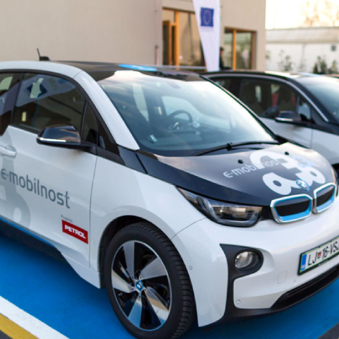 Na sliki je električni avtomobil BMW i3 z oznako »e-mobilnost« in logotipom Petrol je parkiran na označenem mestu za električna vozila. V ozadju so vidni še drugi avtomobili in stavba z evropsko zastavo.