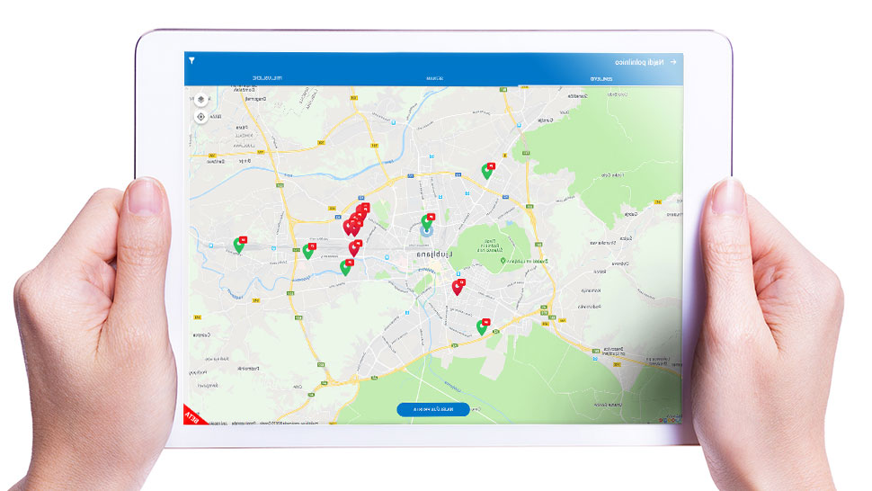 Na sliki sta dve roki, ki držita tablico, na zaslonu katere je prikazan zemljevid z označenimi lokacijami električnih polnilnic. Vmesnik ima modro zgornjo in spodnjo vrstico, na zemljevidu pa so vidni rdeči in zeleni označevalci polnilnic.