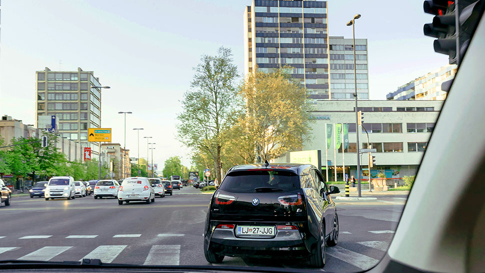 Na sliki je električni avtomobil BMW i3 s črno karoserijo, ki vozi po mestnem križišču. Posnetek je narejen iz notranjosti drugega vozila, v ozadju pa so vidne mestne stavbe, promet in drevesa.