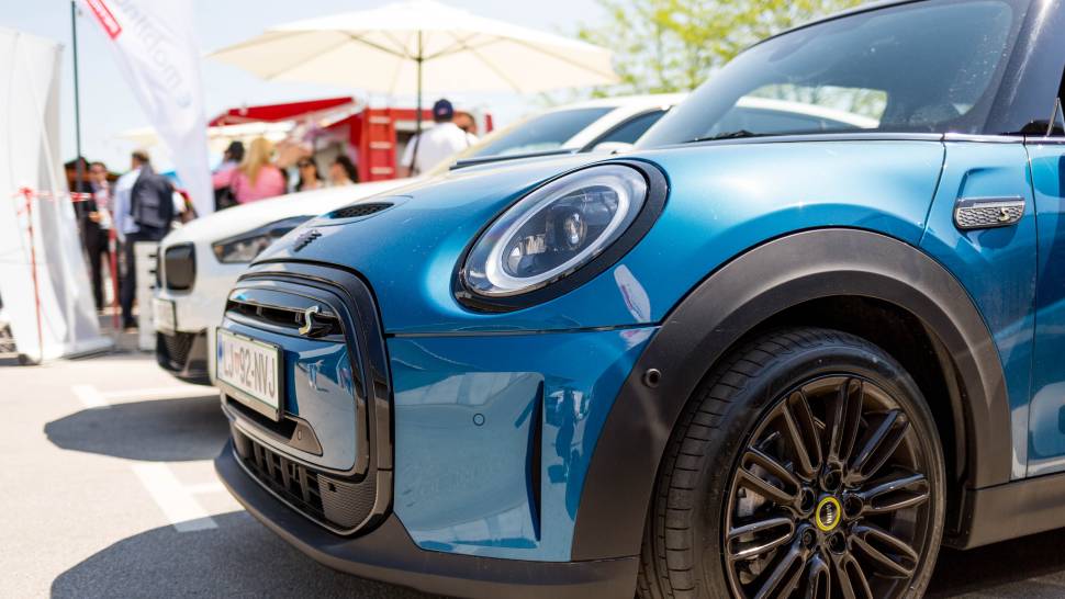 Na sliki je modri električni avtomobil Mini, parkiran na sončen dan ob beli vozilu v ozadju. V ozadju so vidni ljudje, senčniki in zastavice z logotipom Petrol.