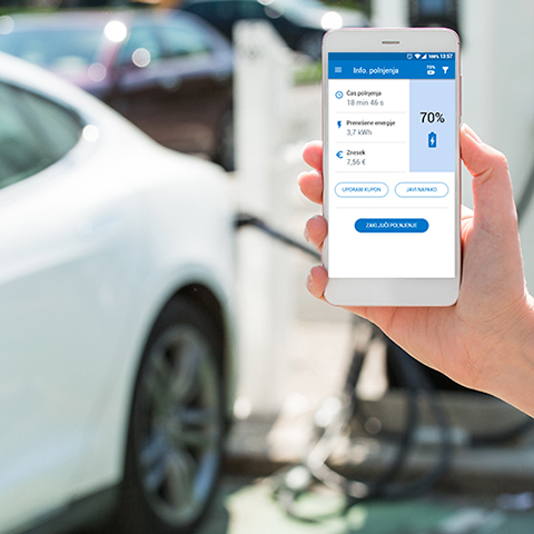 Na sliki je roka osebe, ki drži pametni telefon z odprto aplikacijo OneCharge. Na zaslonu so prikazani podatki o času polnjenja, porabljeni energiji in stanju napolnjenosti baterije. V ozadju je električni avtomobil, priključen na polnilnico.