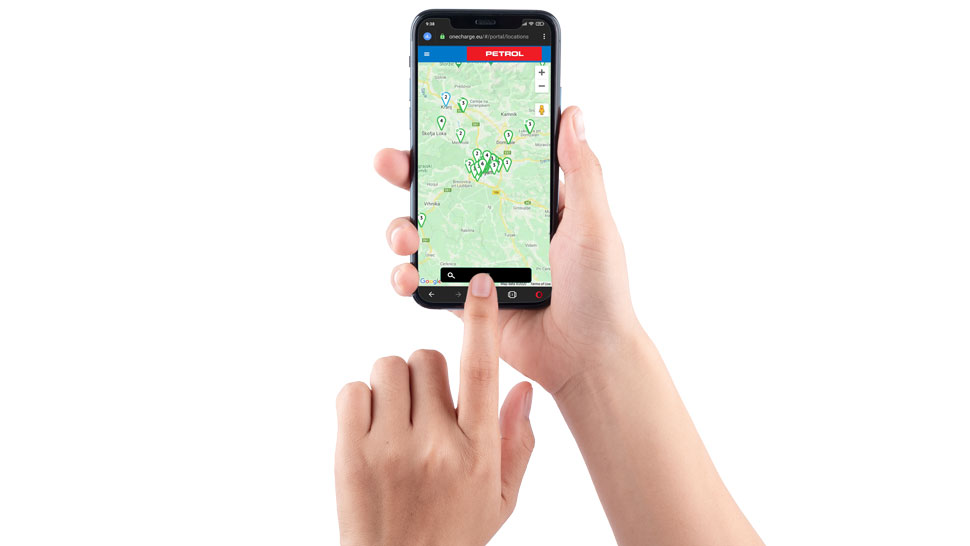 Na sliki sta roki osebe, ki uporabljata pametni telefon z odprto spletno aplikacijo Petrol OneCharge. Na zaslonu je prikazan zemljevid z označenimi lokacijami električnih polnilnic.
